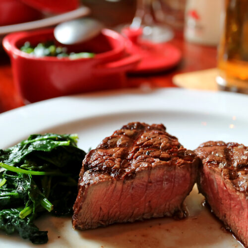 Medium grilled baseball sirloin steak cut in half with sauteed greens on white plate, blurred side dishes and beverage in background
